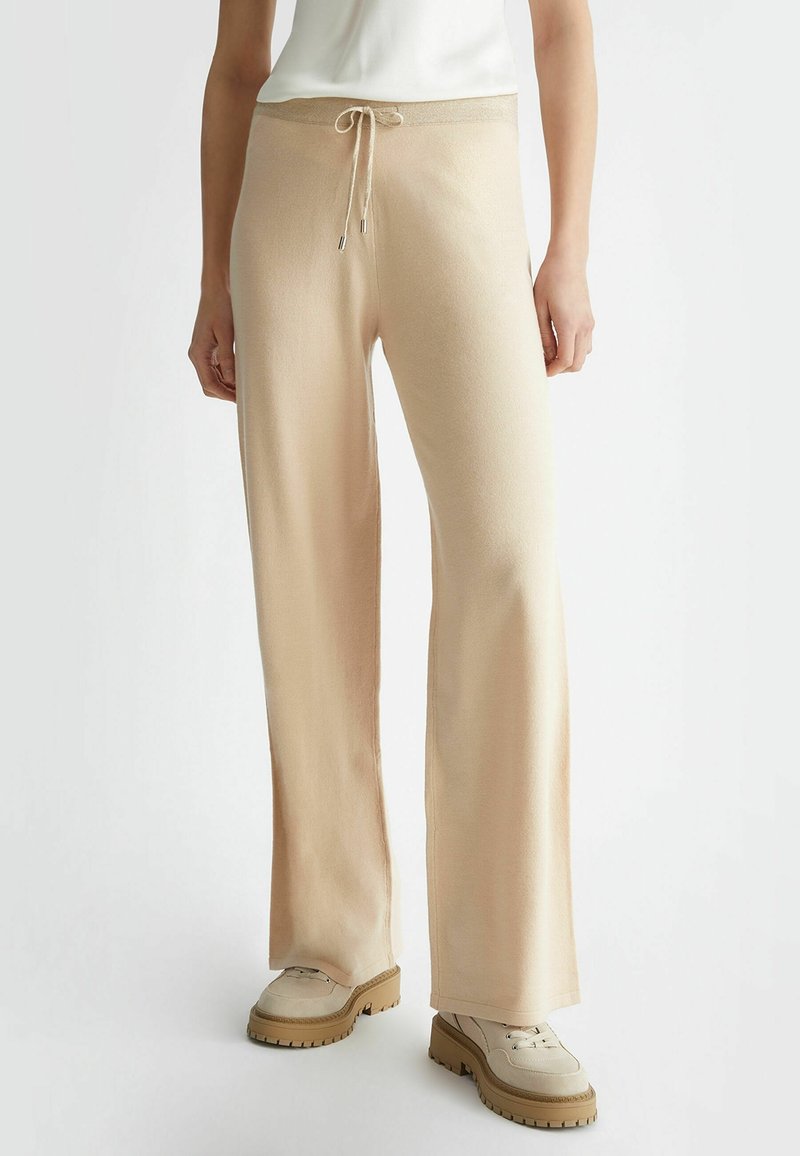 Personne portant un pantalon large en maille beige avec une taille à cordon et des chaussures à semelle épaisse crème sur un fond blanc uni.