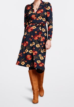 Vrouw draagt een marineblauwe, lange mouwen jurk met een bloemenprint in rood en geel, gecombineerd met kniehoge bruine leren laarzen.