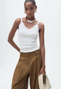 Top bianco a costine con scollo a V, abbinato a pantaloni larghi marroni. La modella tiene una borsa in paglia intrecciata, arricchita da dettagli in argento.