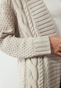 Béžový pletený cardigan s texturovanými kabelkovými a žebrovanými vzory. Má široké rukávy a útulný, silný pletený design.