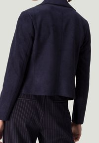 Henkilö, jolla on päällään tumma laivastonsininen takki tiukasti istuvien mustavalkoraitapolvijousten päällä, kuvattuna selkäpuolelta vaaleanharmaata taustaa vasten.