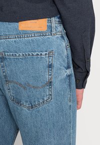 Jeans de denim azul com um acabamento texturizado, apresentando costura nos bolsos traseiros e um patch de couro castanho na cintura com texto em relevo.