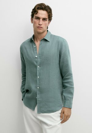 Giovane uomo con indosso una camicia verde chiaro con bottoni e pantaloni bianchi, in piedi rilassato contro un muro bianco.