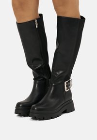 Des bottes noires jusqu'au genou en cuir lisse, dotées d'une fermeture éclair sur le côté et d'un accent de boucle métallique au niveau de la cheville, avec une semelle épaisse.