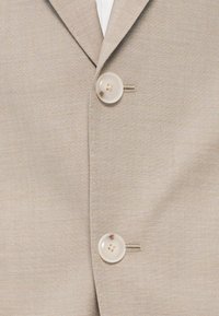 Beige blézer textúrált szövettel, két kerek, világos színű gombbal és egy kis mellzseb részlettel.