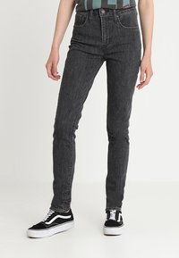 Jeans ajustados de tiro alto en negro, hechos de denim, con un diseño clásico que incluye trabillas para cinturón y estilo de cinco bolsillos, combinados con zapatillas negras.