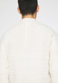 Pull en tricot épais crème, présentant des motifs texturés et des motifs en zigzag. Encolure ronde avec des accents en côtes et des détails en tricot torsadé.