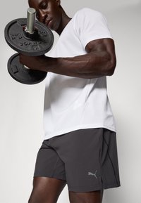 Svart manualholdt i en muskuløs arm, iført en hvit kortermet topp og mørke shorts med Puma-logo. Jevn tekstur, atletisk passform.