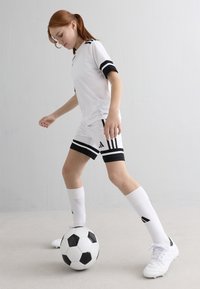 Niño con equipamiento de fútbol en blanco y negro controla un balón de fútbol negro y blanco frente a un fondo blanco liso.
