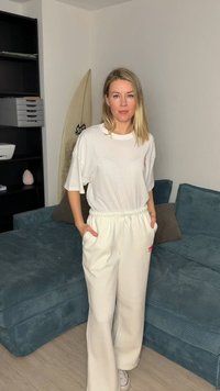 Νεαρή γυναίκα με ξανθά μαλλιά, ντυμένη με λευκό φαρδύ μπλουζάκι και κρεμ παντελόνι, στέκεται σε ένα σαλόνι με τιρκουάζ καναπέ και σανίδα του σερφ.