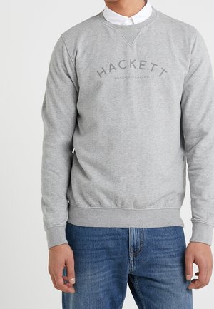 Mężczyzna w jasnoszarej bluzie Hackett London England na białej koszuli i niebieskich dżinsach, stojący na jednolitym tle.