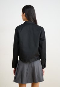 Veste noire courte en tissu lisse avec des manches longues, présentant un col et accentuée de détails en boutons à la taille.
