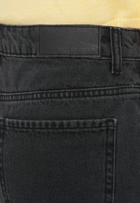 Πίσω μέρος μαύρου τζιν με ορατή ραφή και δερμάτινη ετικέτα μάρκας "Noisy May", φορεμένο με κίτρινο πλεκτό τοπ.