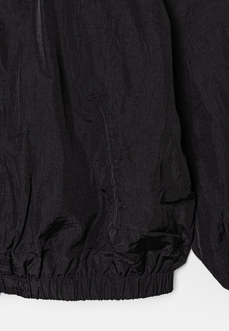 Veste noire en tissu léger et texturé. Comprend un ourlet élastique et une poche sur le côté gauche pour plus de fonctionnalité.