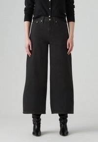 Svarte vide jeans laget av denim, høyt liv med synlige sømmer, to frontlommer og metalliske detaljer; kombinert med knelange støvler.