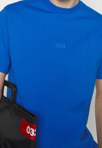 Modré bavlněné tričko s jemným potiskem na přední straně s číslem "424." Černá taška s červenou akcentní etiketou zobrazující "032."