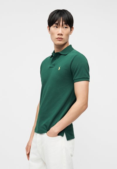 Νεαρός άνδρας με πράσινο κοντομάνικο πόλο πουκάμισο και άσπρο παντελόνι, στέκεται με το ένα χέρι στην τσέπη, κοιτάζοντας προς τα πλάγια, μπροστά σε λευκό φόντο.