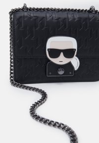 Musta teksturoitu käsilaukku, jossa koristeellinen metallinen kasvokuvio, ketjuhihna ja hopeiset yksityiskohdat. Etupuolella painettu logo.