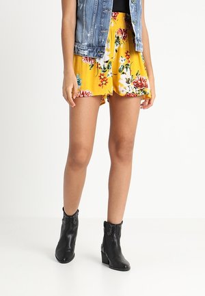 Mujer con pantalones cortos amarillos de flores, una chaqueta vaquera sin mangas y botines negros con tachuelas, de pie sobre un suelo blanco.