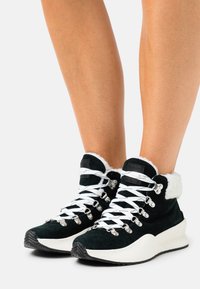 Czarne zamszowe sneakersy typu high-top z białym futrem i białymi sznurówkami. Posiadają metalowe oczka oraz teksturowaną, żłobioną białą podeszwę.