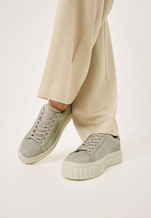 Baskets en cuir vert clair avec de semelles épaisses et texturées et des lacets ton sur ton, présentées avec un pantalon beige, mettant en valeur le design de la chaussure.