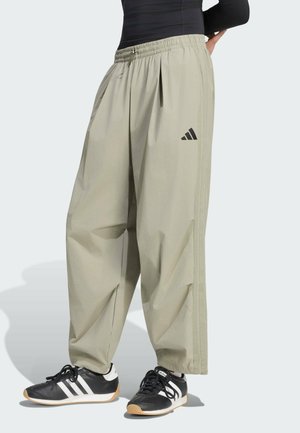 Persona con pantalones beige sueltos con el logo de Adidas y zapatillas negras Adidas con rayas blancas, de pie contra un fondo liso.