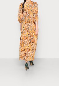 Żółto-pomarańczowa maxi sukienka z nadrukiem paisley, z bufkami na rękawach i cinczonym w talii. W zestawie z zielonymi szpilkami.