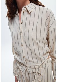 Camicia beige con bottoni, colletto, caratterizzata da righe verticali scure, vestibilità ampia e dettagli arricciati in vita con accentuazioni in cravatta.