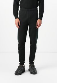 Black sweatpants with an elastic waistband, featuring green side panels, a tapered fit, and ribbed ankle cuffs. Paired with black sneakers.