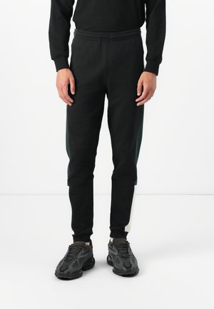 Pantaloni da ginnastica neri con vita elasticizzata, dotati di pannelli laterali verdi, vestibilità affusolata e polsini a coste sulle caviglie. Abbinati a sneakers nere.