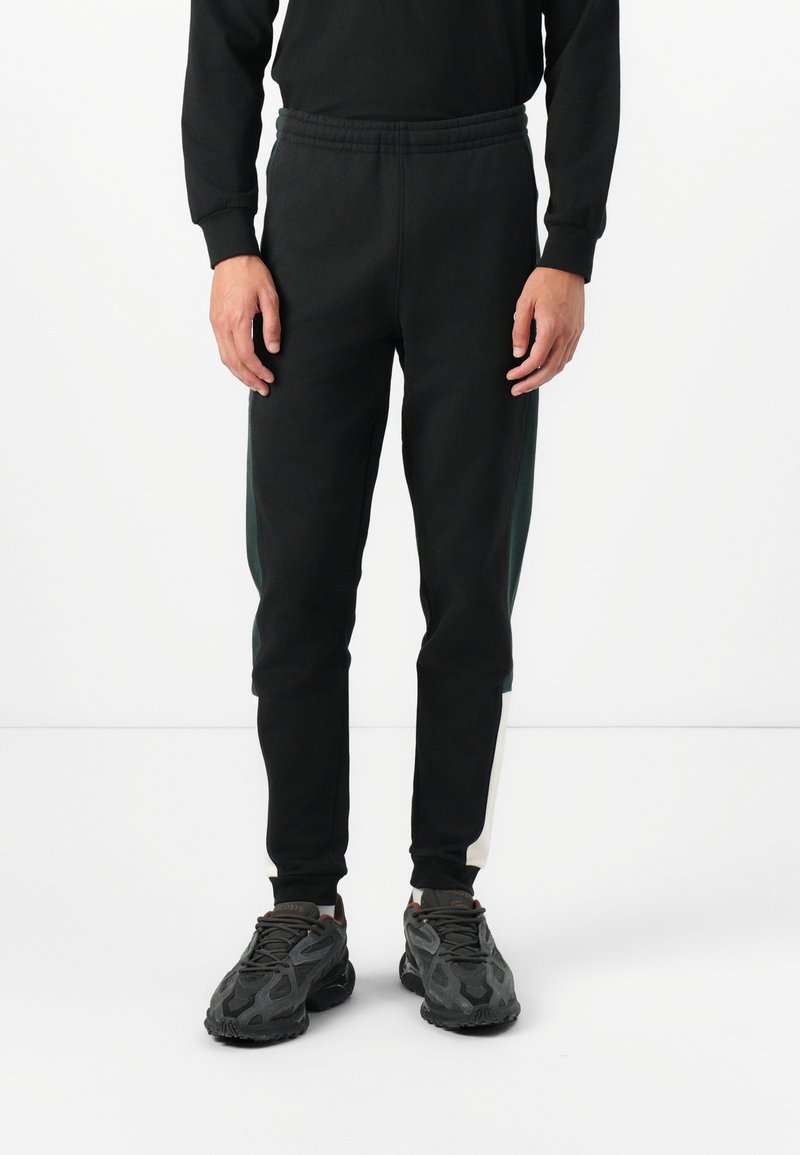 Black sweatpants with an elastic waistband, featuring green side panels, a tapered fit, and ribbed ankle cuffs. Paired with black sneakers.