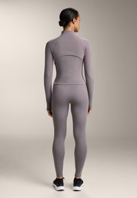 Svetlofialová dlhá rukávová blúzka s vysokým golierom, priliehavý strih, bezšvové akcenty; kombinovaná s padnúcimi legínami a čiernymi teniskami.