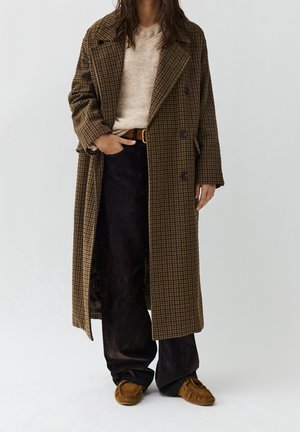 Personne portant un long manteau marron à carreaux, un pull beige, un pantalon foncé, une ceinture marron et des chaussures style mocassin beige, debout avec une main dans une poche.