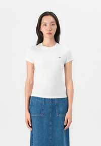 Hvid tætsiddende t-shirt med korte ærmer, rund hals og lille logo detaljer på brystet, parret med en denimnederdel. Glat stoftekstur.