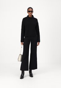 Maglione nero a costine con collo alto, abbinato a pantaloni neri a gamba larga e stivaletti neri. Si tiene in mano una piccola borsa di colore chiaro.