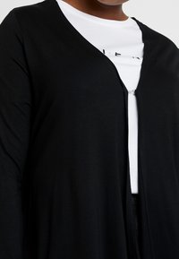 Osoba w czarnym kardiganie zapinanym na pojedynczy zamek, nosząca białą koszulę z częściowym czarnym tekstem.