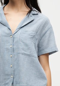 Femme portant une chemise à manches courtes à petits carreaux bleus et blancs, avec une poche poitrine et un col.