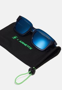 Námornícke modré slnečné okuliare s gradientnými modrými šošovkami v čiernom puzdre so zeleným brandingom "ARNETTE" a zatahovacou šnúrkou na uzatvorenie.
