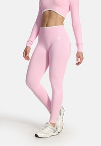 Femme portant des leggings roses taille haute et un top long à manches longues court avec des baskets blanches, debout devant un fond blanc.