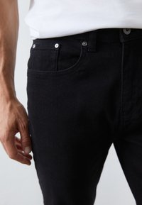 Vaqueros de mezclilla negros con un corte ajustado, que presentan un diseño clásico de cinco bolsillos y botones de metal plateado. Se suministran con una camiseta blanca lisa.