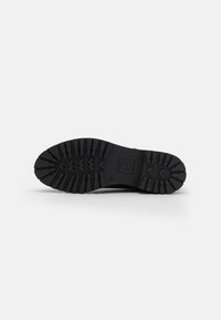 Semelle de chaussure en caoutchouc noir avec des dessins en zigzag, des profondeurs marquées et un logo de marque rectangulaire. La surface est texturée et durable.