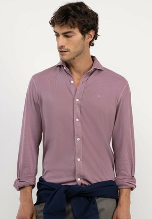 Hombre con cabello rizado y oscuro, vestido con una camisa color malva con botones y un suéter azul marino atado alrededor de su cintura, mirando hacia su izquierda.