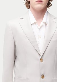 Blazer gris clair en tissu texturé, avec une coupe à simple boutonnage, des revers crantés et deux boutons. Chemise blanche en dessous.