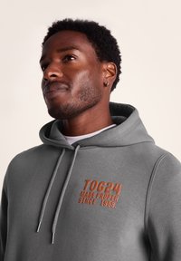 Gray hoodie featuring a kangaroo pocket and drawstring hood. Embroidered logo in brown on the chest, with silver-tone hardware on the drawstrings.