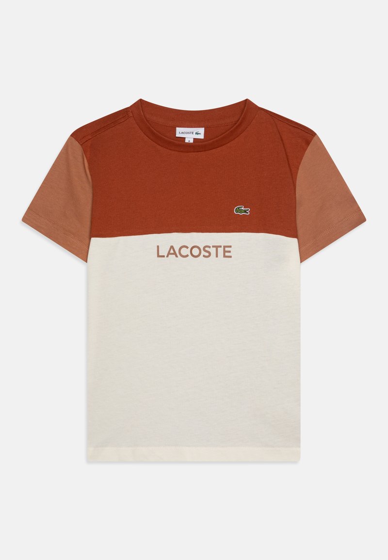 Tričko s barevnými bloky, s horní částí v barvě rezavé a spodní částí v krémové barvě. Obsahuje hnědý nápis "LACOSTE" a malý zelený krokodýl jako logo. Materiál: bavlna.