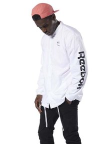 Giacca leggera bianca con bottoni a scatto e logo sul petto. Le maniche lunghe presentano la scritta "Reebok" in lettere nere in grassetto. Pantaloni neri.