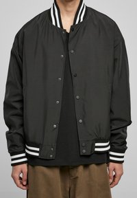 Czarna kurtka bomber wykonana z gładkiego materiału z prążkowanym kołnierzem. Posiada białe paski na mankietach oraz zapięcie na zatrzaski z przodu.