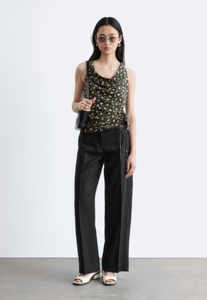 Kobieta ubrana w czarny, kwiecisty top bez rękawów, czarne spodnie o szerokich nogawkach, białe buty na obcasie z odkrytymi palcami, okulary przeciwsłoneczne i nosząca czarną torebkę na ramię.