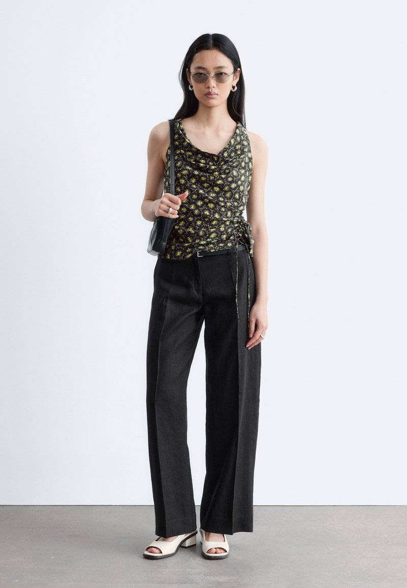 Femme portant un top sans manches noir à fleurs, un pantalon noir à jambes larges, des talons blancs ouverts, des lunettes de soleil et un sac à épaule noir.