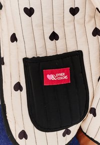 Bundi s prešívaním, ktorý má krémovú tkaninu s čiernymi srdcovými vzormi a vertikálnymi pruhmi. Obsahuje čiernu kapsu s červeným štítkom.